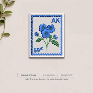 May include: A rectangular embroidered patch designed like a postage stamp with a white background and a blue scalloped border. It features blue forget-me-not flowers with green stems and leaves, the letters AK, and the text 59 cents in blue thread.