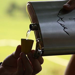 Könnte beinhalten: Eine Edelstahl-Flachmann mit Berg- und Hüttendesign gießt Flüssigkeit in ein kleines gelbes Schnapsglas. Der Flachmann wird von einer Person gehalten, mit einem verschwommenen grünen Hintergrund.