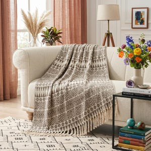 May include: A cream-coloured sofa with a patterned throw blanket. The throw has a geometric design in brown and cream, with a fringed edge. The room also features a rug, lamp, and a vase of flowers.