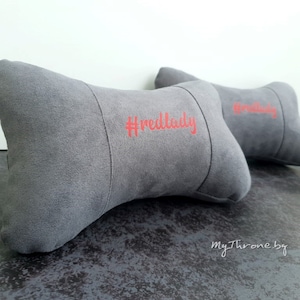 May include: Two gray car neck pillows with the text "#redlady" embroidered in red on one pillow.