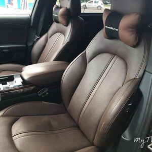 May include: Two brown leather car seats with headrests. The headrests have brown and black quilted neck pillows with the word "quattro" printed on the right pillow.