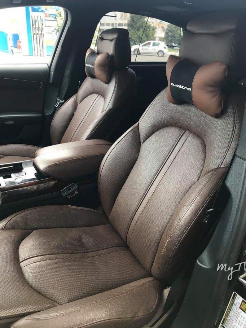 May include: Two brown leather car seats with headrests. The headrests have brown and black quilted neck pillows with the word "quattro" printed on the right pillow.