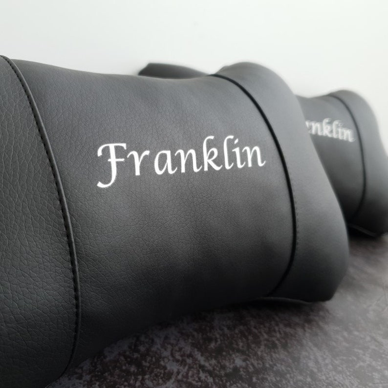 May include: Two black leather car headrests with white embroidery. The headrest on the left has the name "Franklin" embroidered on it.