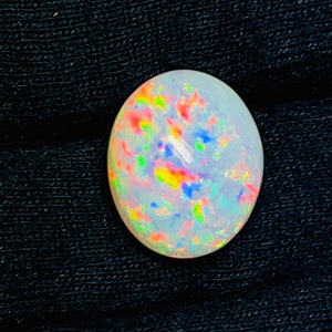 Op de afbeelding: Ovale opaal edelsteen met een melkachtige witte basis en levendig intern kleurenspel. De edelsteen toont flitsen van rood, geel, blauw en groen, tegen een donkere, getextureerde achtergrond. Ideaal voor het maken van sieraden.