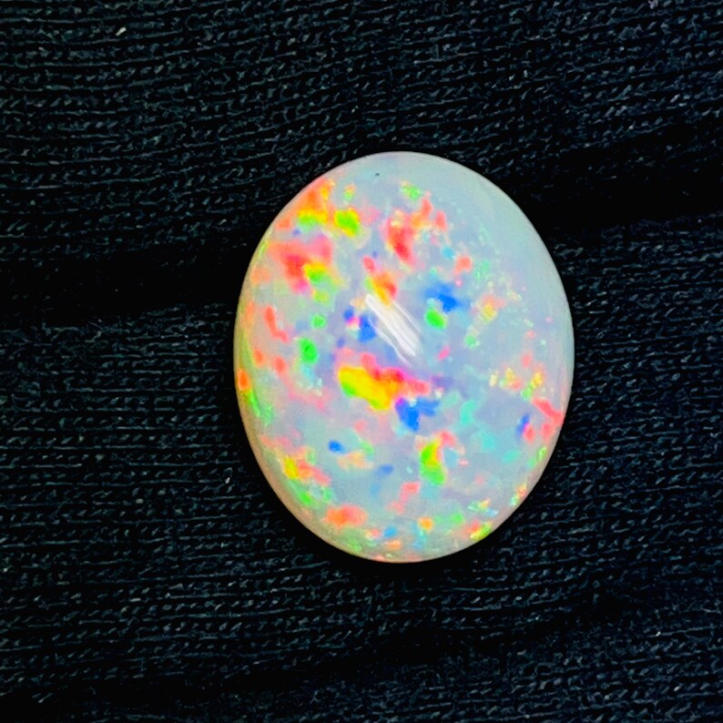 Op de afbeelding: Ovale opaal edelsteen met een melkachtige witte basis en levendig intern kleurenspel. De edelsteen toont flitsen van rood, geel, blauw en groen, tegen een donkere, getextureerde achtergrond. Ideaal voor het maken van sieraden.