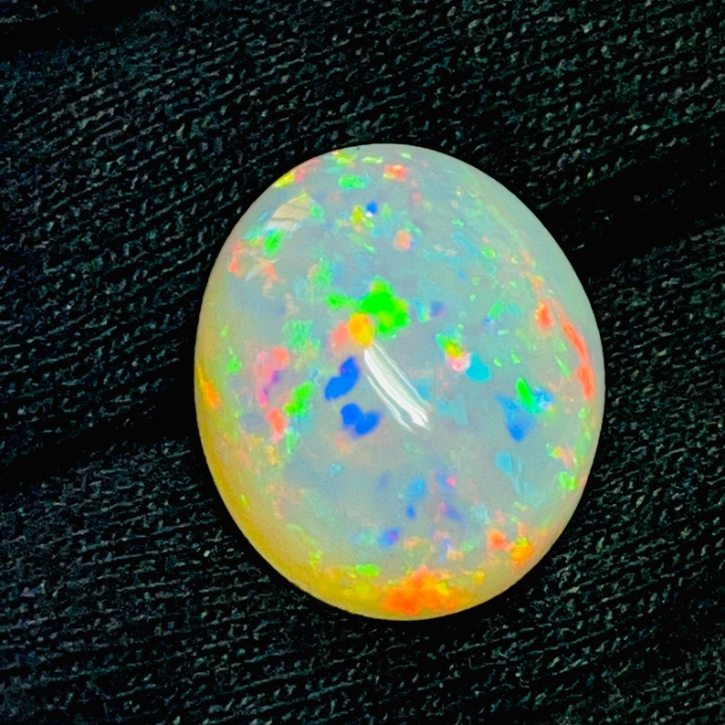 Op de afbeelding: Een gepolijste, ovaalvormige opaal edelsteen. De opaal heeft een melkachtige witte basiskleur met iriserende flitsen van blauw, groen, oranje en geel. De edelsteen is geplaatst tegen een donkere, getextureerde achtergrond.