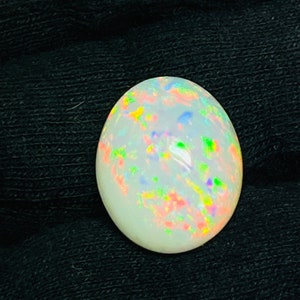 Op de afbeelding: Ovale opaal edelsteen met een melkachtige witte basis en levendige kleurflitsen. De opaal toont een kleurenspel effect, met vlekken van rood, groen, blauw en geel. De edelsteen is geplaatst tegen een donkere achtergrond, wat de iriserende kwaliteiten benadrukt.