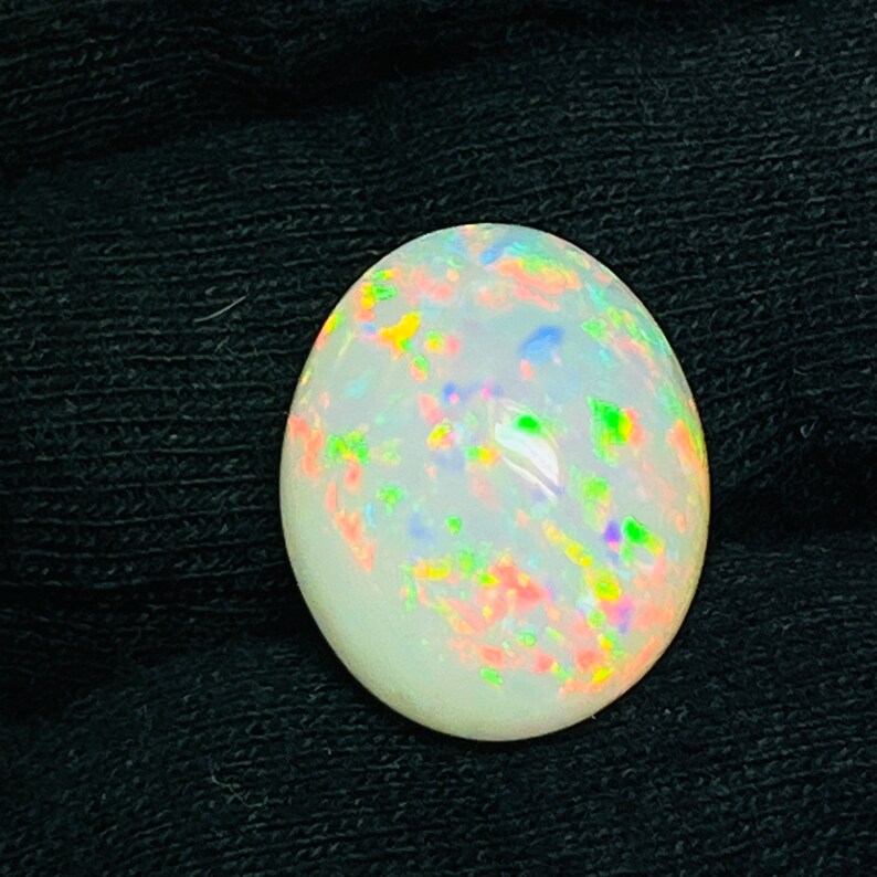 Op de afbeelding: Ovale opaal edelsteen met een melkachtige witte basis en levendige kleurflitsen. De opaal toont een kleurenspel effect, met vlekken van rood, groen, blauw en geel. De edelsteen is geplaatst tegen een donkere achtergrond, wat de iriserende kwaliteiten benadrukt.