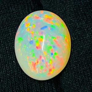 Op de afbeelding: Ovale opaal edelsteen met een melkachtige witte basis en levendige kleurflitsen. De edelsteen vertoont een kleurenspel-effect met vlekken van rood, geel, groen en blauw. De opaal is geplaatst tegen een donkere achtergrond.