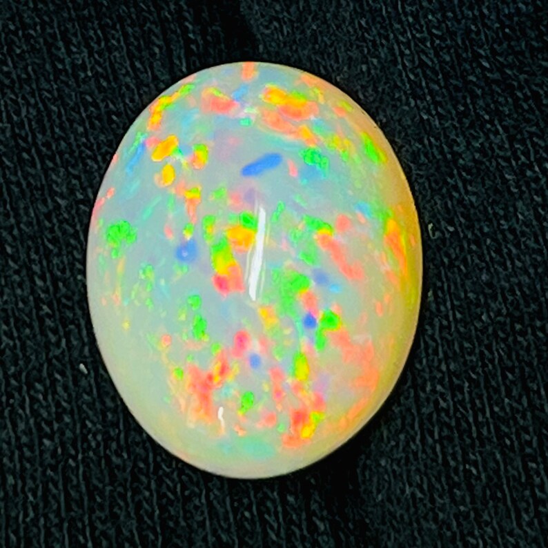 Op de afbeelding: Ovale opaal edelsteen met een melkachtige witte basis en levendige kleurflitsen. De edelsteen vertoont een kleurenspel-effect met vlekken van rood, geel, groen en blauw. De opaal is geplaatst tegen een donkere achtergrond.