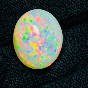 Op de afbeelding: Een gepolijste, ovaalvormige opaal edelsteen. De opaal heeft een melkachtige witte basiskleur met levendige kleurflitsen, waaronder roze, groen, geel en blauw. De edelsteen is geplaatst tegen een donkere, getextureerde achtergrond.