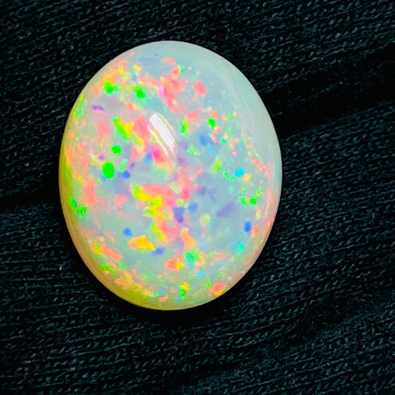 Op de afbeelding: Een gepolijste, ovaalvormige opaal edelsteen. De opaal heeft een melkachtige witte basiskleur met levendige kleurflitsen, waaronder roze, groen, geel en blauw. De edelsteen is geplaatst tegen een donkere, getextureerde achtergrond.