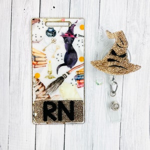 May include: A gold glitter witch hat with a black cat and a broom on a white background with the letters RN. The badge holder is made of clear plastic and has a gold glitter witch hat charm.