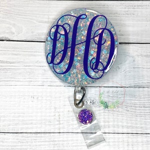 May include: A white glitter badge holder with a blue and white glitter circle featuring the monogram "DHD" in a dark blue script font. The badge holder has a clear plastic strap and a purple glitter circle attached to the strap.