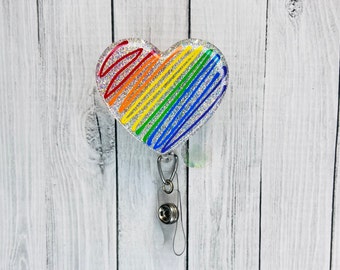 May include: A heart-shaped badge reel with a rainbow scribble design. The badge reel has a clear, retractable cord and a silver-colored clip. The heart is outlined with silver glitter and has a clear, glossy finish.