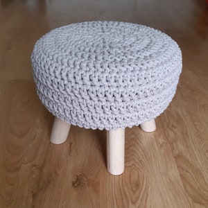 Könnte beinhalten: Ein grauer Häkelhocker mit Holzbeinen. Der Hocker ist rund und hat eine strukturierte Oberfläche.