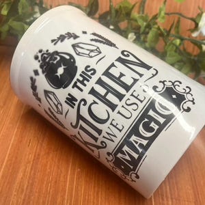 May include: A white ceramic kitchen utensil holder with black graphics and text. The text reads "IN THIS KITCHEN WE USE MAGIC" with illustrations of a cauldron, crystals, and herbs. The holder is on a wooden surface.