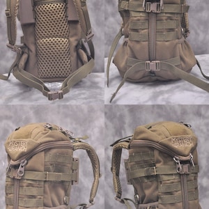 May include: A tan backpack with multiple straps, buckles, and a mesh back panel. The top of the pack has the text "MYSTERY RANCH" in white. The backpack has adjustable shoulder straps and a zippered main compartment.