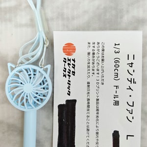 May include: A light blue handheld fan with a cat-shaped design. The fan has a white and blue blade and a white string attached to it. The fan is 60 cm long and is designed for 1/3 scale dolls. The packaging includes the text "ニャンディ・ファン L" and "1/3 (60cm) ドール用".