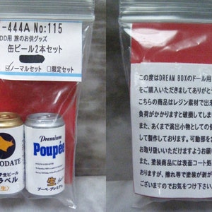 May include: Two miniature cans of beer, one gold and one silver, with Japanese text. The cans are labelled "Hakodate" and "Premium Poupée". The cans are packaged in a clear plastic bag with a red background.
