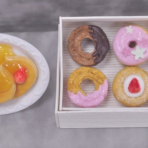 May include: A white box containing six assorted donuts. The donuts are decorated with pink icing, chocolate icing, and white sprinkles. There is also a plate of three mini pancakes topped with strawberry and syrup.