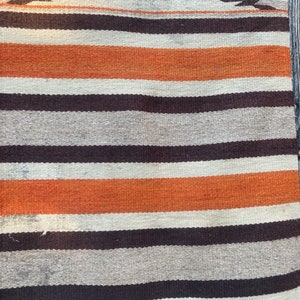 May include: A striped woven rug with orange, brown, and white stripes. The rug is hanging on a wooden wall.