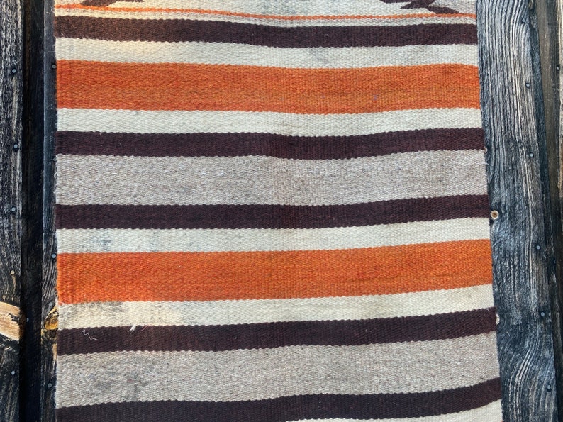 May include: A striped woven rug with orange, brown, and white stripes. The rug is hanging on a wooden wall.