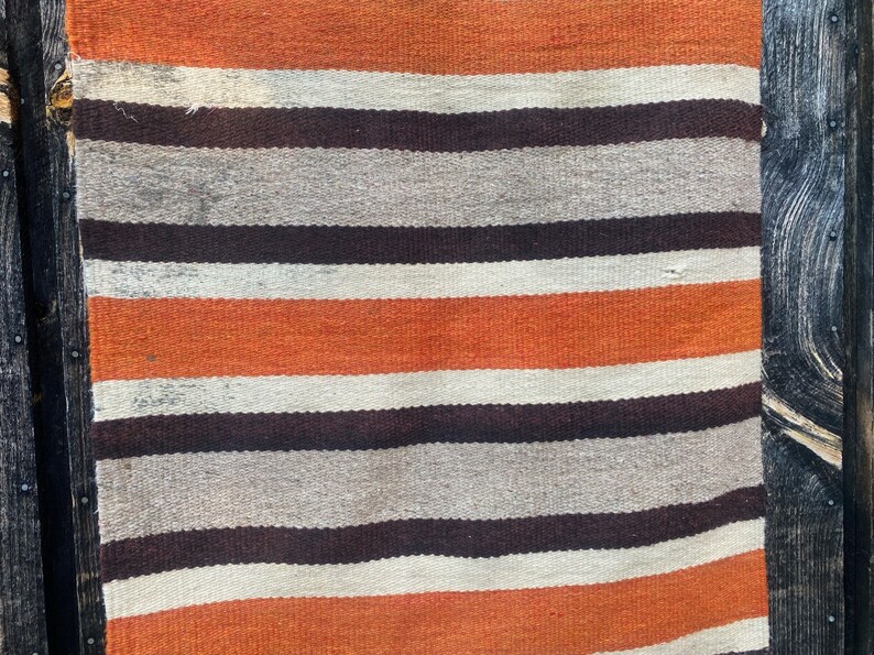 May include: A striped woven rug with orange, white, and brown stripes. The rug is hanging on a dark wood wall.