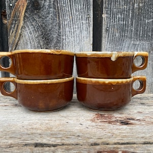Può includere: Quattro ciotole da zuppa in ceramica marrone con manici sono impilate su una superficie di legno. Le ciotole hanno una forma arrotondata con un bordo e un manico di colore più chiaro. Lo sfondo è un muro di legno stagionato.