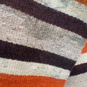 May include: Close-up of a woven fabric with stripes of brown, white, and orange. The fabric has a rough texture and is likely made of wool.