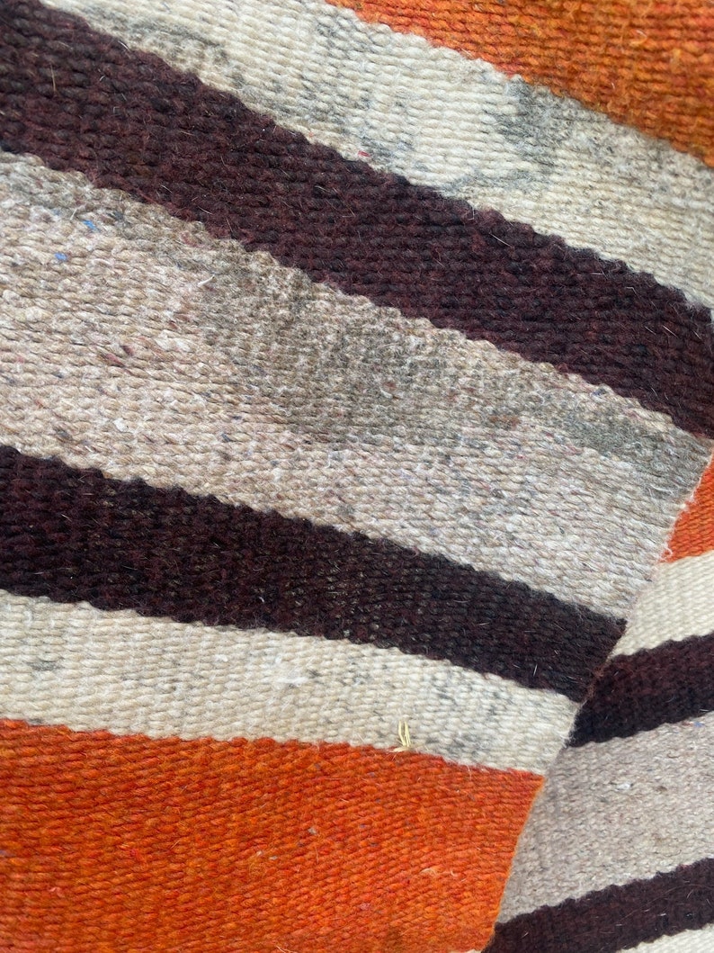 May include: Close-up of a woven fabric with stripes of brown, white, and orange. The fabric has a rough texture and is likely made of wool.