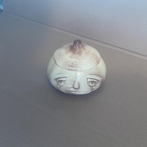 May include: A ceramic onion-shaped jar with a lid, featuring a light cream glaze with brown shading. The jar has a sculpted face with sad, teary eyes and a downturned mouth. The word ONION is embossed on the lid, which has a small brown stem top.
