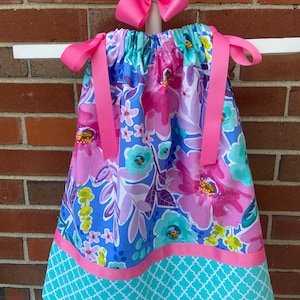 May include: A blue and pink floral print dress with a pink ribbon sash and a matching pink bow. The dress has a turquoise and white geometric pattern at the bottom.
