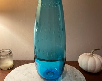 Könnte beinhalten: Eine hohe, türkisblaue Glasvase mit strukturierter Oberfläche. Die Vase hat einen abgerundeten Boden und eine schmale Öffnung.