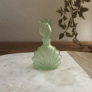 Könnte beinhalten: Eine gefrostete grüne Glasparfümflasche mit einem tulpenförmigen Stopfen. Die Flasche hat ein Muschel-Design und steht auf einer weißen Marmorfläche.