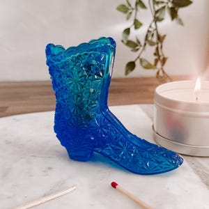 May include: A blue glass boot-shaped vase with a textured surface. The vase is sitting on a white marble surface with two wooden matches.