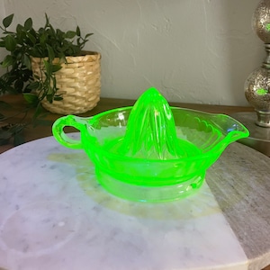 Peut inclure: Un presse-agrumes en verre vert vif avec un sommet cannelé et deux poignées. Le presse-agrumes est posé sur une surface en marbre blanc.