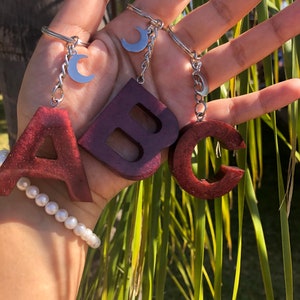 May include: Three wooden keychains shaped like the letters A, B, and C. Each keychain has a silver crescent moon charm attached to a silver chain.