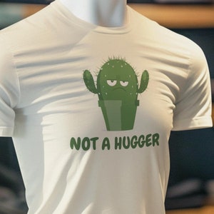 May include: White t-shirt featuring a green cactus graphic and the text "NOT A HUGGER". The t-shirt is displayed on a mannequin in a clothing store.