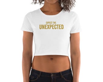 May include: White cropped t-shirt with short sleeves and the text "Expect the Unexpected" in gold lettering.