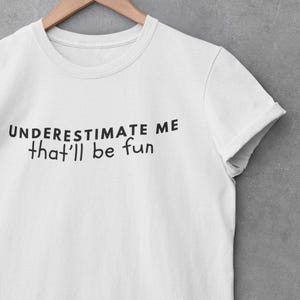 May include: White t-shirt with black text that reads "UNDERESTIMATE ME that'll be fun".