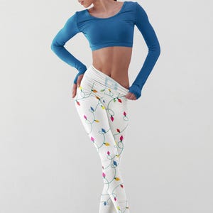 May include: A woman wearing a blue long-sleeved crop top and white leggings with a colourful string of lights pattern. The leggings are tight-fitting and show off her legs. The woman is standing on a white background.