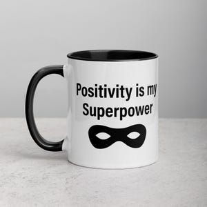 May include: White ceramic mug with a black interior and handle. The mug features the text "Positivity is my Superpower" in black, along with a black superhero mask graphic. The mug is designed for hot or cold drinks.