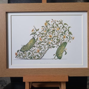 May include: A framed print of a watercolour illustration of a high-heeled shoe with a cucumber growing out of it. The shoe is made of vines and leaves, and the cucumber is green with yellow flowers. The print is signed "Eleanor Shiels".