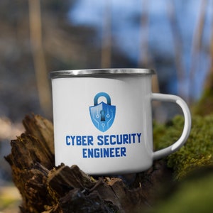 Könnte beinhalten: Eine weiße Emailletasse mit einem blauen und weißen Logo eines Vorhängeschlosses mit einem Schlüsselloch und einem Schaltkreisdesign. Der Text "Cyber Security Engineer" ist unter dem Logo aufgedruckt.