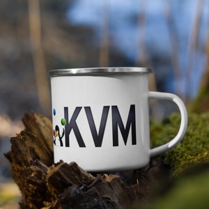 May include: White enamel mug with a black KVM logo and a small penguin illustration. The mug is sitting on a piece of weathered wood.