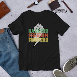Könnte beinhalten: Schwarzes T-Shirt mit einer weißen Faustgrafik und dem Text "Hackers Freedom Fighters" in Grün, Rot und Gelb.