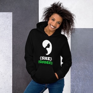 May include: Black hoodie with a white silhouette of a woman's head and the text "{SHE} [CODES];" in white and green.