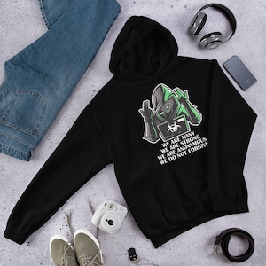 May include: Black hooded sweatshirt featuring a green graphic of a hooded figure holding a laptop with a biohazard symbol. The text "We are many, We are strong, We are anonymous, We do not forgive" is printed on the sweatshirt.