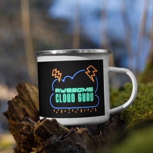 Könnte beinhalten: Eine weiße Emailletasse mit schwarzem Hintergrund und einer grünen Wolke mit dem Text "AWESOME CLOUD GURU" in neonorange und grüner Schrift. Auf beiden Seiten der Wolke befinden sich zwei orangefarbene Blitze.